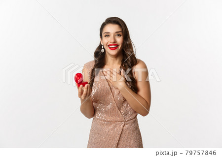 Image of beautiful woman in glamour dress receiving engagement ring, looking surprised and happy at camera, being proposed, standing over white background 79757846