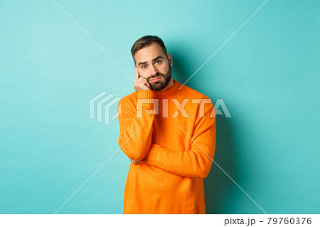 Sad and gloomy man with beard, looking indesicive and sad, thinking about something and sulking, standing over turquoise background Sad and gloomy man with beard, looking indesicive and sad, thinking about something and sulking, standing over turquoise background 79760376