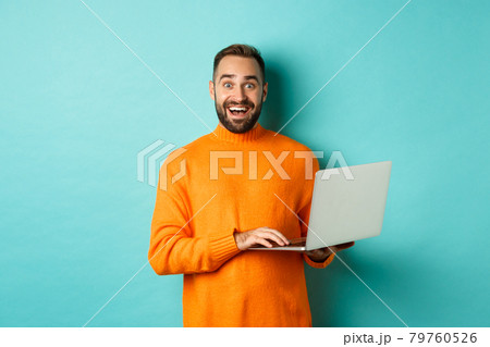 Excited smiling man working on laptop, staring at camera happy, standing in orange sweater against turquoise background Excited smiling man working on laptop, staring at camera happy, standing in orange sweater against turquoise background 79760526