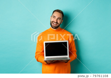 Happy caucasian man thinking and showing laptop screen, smiling pleased while pondering, standing in orange sweater against light blue background Happy caucasian man thinking and showing laptop screen, smiling pleased while pondering, standing in orange sweater against light blue background 79761108