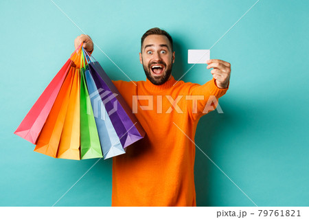 Happy aduly man showing credit card and shopping bags, standing against turquoise background Happy aduly man showing credit card and shopping bags, standing against turquoise background 79761821