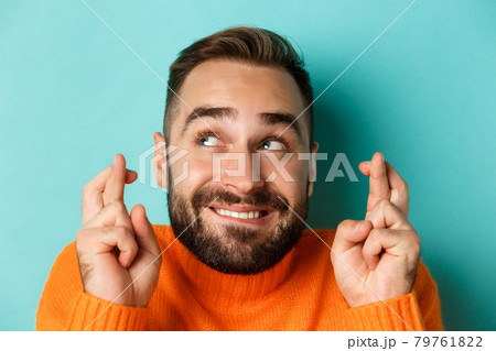 Headshot of hopeful bearded man making a wish, smiling and holding fingers crossed for good luck, standing over light blue background 79761822