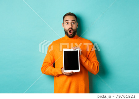 Technology. Surprised bearded man drop jaw, staring at camera and showing digital tablet screen, standing againt light blue background Technology. Surprised bearded man drop jaw, staring at camera and showing digital tablet screen, standing againt light blue background 79761823