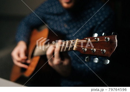 Close-up of unrecognizable guitarist singer male playing acoustic guitar sitting on armchair in dark living room, selective focus. Close-up of unrecognizable guitarist singer male playing acoustic guitar sitting on armchair in dark living room, selective focus. 79764596