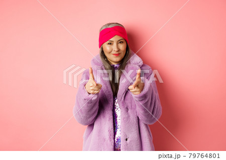 Fashion and shopping concept. Cool asian senior woman in stylish fake fur coat pointing fingers at camera, asking you to check out promo offer, standing over pink background 79764801