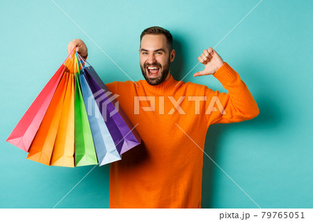 Handsome bearded man in orange sweater, buying gifts, pointing at himself and showing shopping bags, standing over blue background Handsome bearded man in orange sweater, buying gifts, pointing at himself and showing shopping bags, standing over blue background 79765051