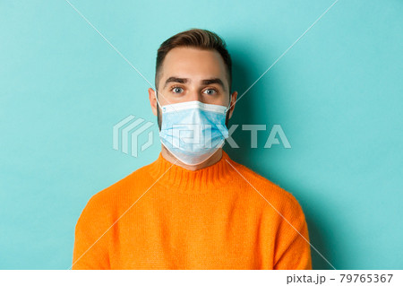 Covid-19, social distancing and quarantine concept. Close-up of young man in face mask staring at camera, standing over turquoise background Covid-19, social distancing and quarantine concept. Close-up of young man in face mask staring at camera, standing over turquoise background 79765367