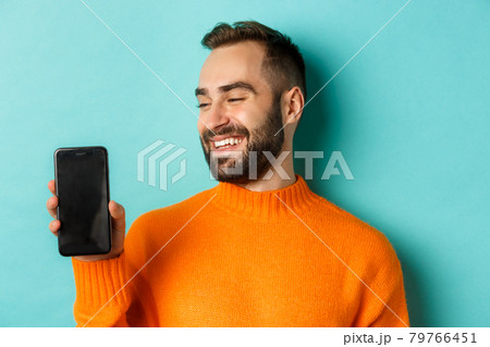 Close-up of young bearded man showing phone screen and looking satisfied, wearing orange sweater, standing against studio background 79766451