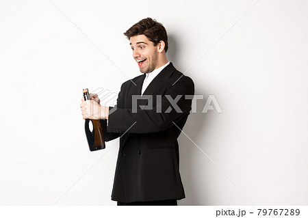 Portrait of attractive man in black suit, winking at camera and opening bottle of champagne, celebrating new year, standing against white background Portrait of attractive man in black suit, winking at camera and opening bottle of champagne, celebrating new year, standing against white background 79767289