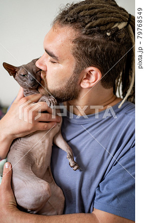 a man with dreadlocks and a beard kisses a purebred bald cat.  79768549