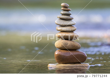 Close-up abstract image of wet rough natural brown uneven different sizes and forms stones balanced like pyramid pile landmark in shallow water on blurred blue-green misty copy space background. 79771244