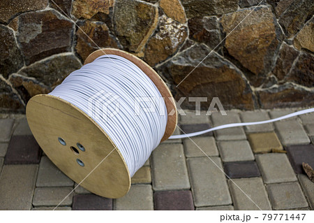 Big roll of white industrial electrical cable on large wooden reel outdoors on sunny pavement on stone fence background. Professional construction site cable, reliable wiring and durability. Big roll of white industrial electrical cable on large wooden reel outdoors on sunny pavement on stone fence background. Professional construction site cable, reliable wiring and durability. 79771447