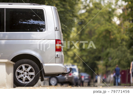 Side view detail of white passenger medium size luxury minibus van parked on summer city street pavement with blurred silhouettes of pedestrians on green trees bokeh copy space background. 79771456
