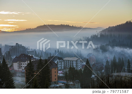 Resort village houses buildings on background of foggy blue mountain hills covered with dense misty spruce forest under bright pink sky at sunrise. Mountain landscape at dawn. 79771587