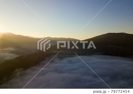 Aerial view of vibrant sunrise over white dense fog with distant dark silhouettes of mountain hills on horizon. 79772454