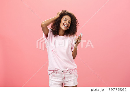Portrait of carefree stylish modern dark-skinned young girl using smartphone hanging around joyfully touching hair and gazing with broad smile at camera holding cellphone posing against pink wall 79776362