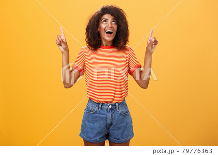 Fascinated impressed and amused good-looking charming African-American woman with afro hairstyle in trendy t-shirt and denim shorts pointing and looking up with interested look over orange wall 79776363