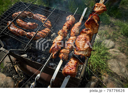 Barbeque meat and sausages or bratwurst on a grill grate in backyard. Man preparing shashlik or shish kebab over charcoal. Grilled meat on metal skewer outdoor. BBQ party or picnic food. Close up shot 79776626