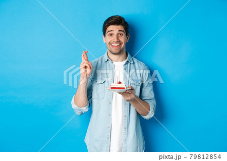 Hopeful man holding birthday cake and cross fingers, making wish, standing over blue background Hopeful man holding birthday cake and cross fingers, making wish, standing over blue background 79812854