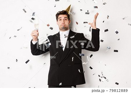 Handsome party guy in black suit having fun, celebrating new year, blowing whistle and drinking champagne while confetti falling, wishing happy holidays, standing against white background 79812889