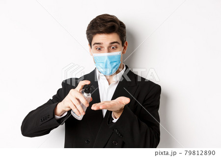 Concept of covid-19, business and social distancing. Image of handsome businessman in trendy suit and medical mask, cleaning hands with hand sanitizer, standing over white background Concept of covid-19, business and social distancing. Image of handsome businessman in trendy suit and medical mask, cleaning hands with hand sanitizer, standing over white background 79812890