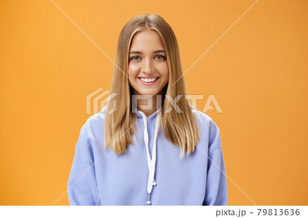 Waist-up shot of pleasant kind and happy friendly-looking young fair-haired girl in comfortable hoodie smiling cheerfully at camera with delighted carefree look posing against orange background 79813636