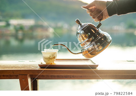 Hand holding vintage teapot and pouring hot tea to cup on wood table against lake view background at coffee shop in the morning sunrise, Ban Rak Thai village, Mae Hong Son province, Thailand 79822584