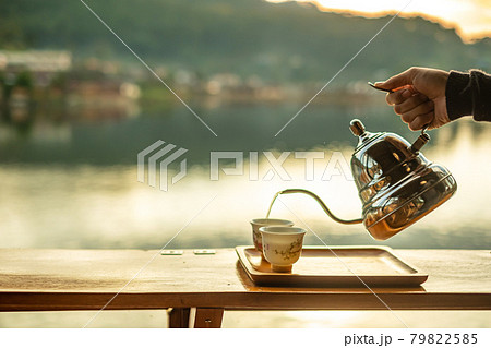 Hand holding vintage teapot and pouring hot tea to cup on wood table against lake view background at coffee shop in the morning sunrise, Ban Rak Thai village, Mae Hong Son province, Thailand 79822585