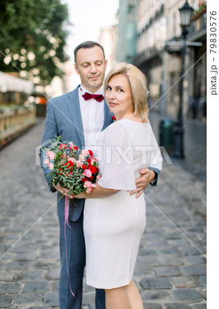 Happy middle aged Caucasian couple, posing with flower bouquet outside in beautiful old city street 79830576