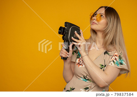 Cheerful young woman holding movie camera and posing against yellow background 79837584
