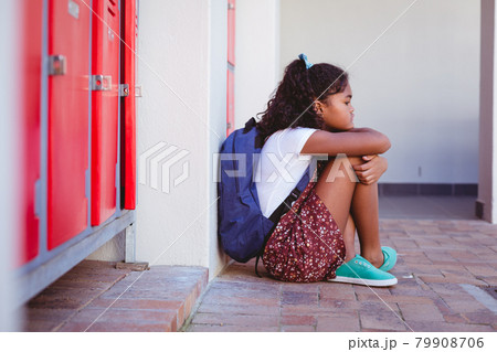 Unhappy african american schoolgirl sitting by lockers in school corridor with schoolbag Unhappy african american schoolgirl sitting by lockers in school corridor with schoolbag 79908706