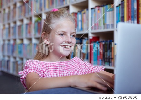 Smiling caucasian schoolgirl at desk in school library using laptop Smiling caucasian schoolgirl at desk in school library using laptop 79908800
