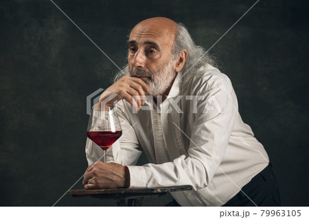 Elderly gray-haired man, medieval hystorical person, actor drinking wine isolated on dark vintage background. Retro style, comparison of eras concept. Elderly gray-haired man, medieval hystorical person, actor drinking wine isolated on dark vintage background. Retro style, comparison of eras concept. 79963105