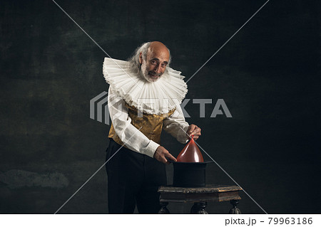 Elderly gray-haired man, actor or magician isolated on dark vintage background. Retro style, comparison of eras concept. Elderly gray-haired man, actor or magician isolated on dark vintage background. Retro style, comparison of eras concept. 79963186