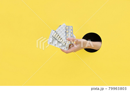 A female hand holds a blister with pills on a yellow background. Hand in a paper hole. Healthcare concept. A female hand holds a blister with pills on a yellow background. Hand in a paper hole. Healthcare concept. 79963803