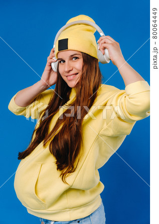 A pregnant girl in a yellow jacket and headphones stands on a blue background 80064449