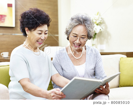 two senior asian women sitting on couch reading a book together 80085010