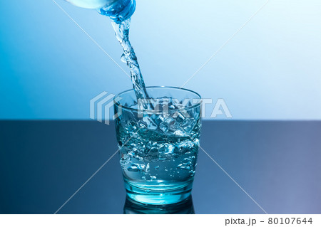 In a glass standing on a black table pour clean water from a bottle. Blue glass with flowing water standing on a black table on a blue background In a glass standing on a black table pour clean water from a bottle. Blue glass with flowing water standing on a black table on a blue background 80107644