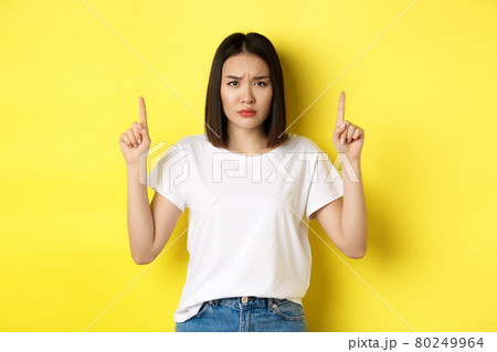 Beauty and fashion concept. Beautiful asian woman in white t-shirt pointing fingers up, standing over yellow background Beauty and fashion concept. Beautiful asian woman in white t-shirt pointing fingers up, standing over yellow background 80249964