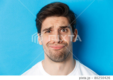 Head shot of skeptical and disappointed man, grimacing complicated, standing against blue background 80250426