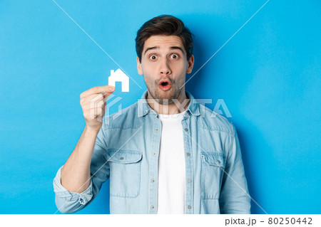 Real estate concept. Surprised guy showing small paper house model, looking amazed, standing against blue background 80250442