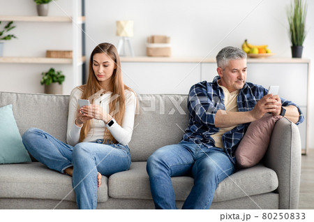 Mature couple using mobile devices, sitting apart on sofa, not paying attention to each other, playing online games Mature couple using mobile devices, sitting apart on sofa, not paying attention to each other, playing online games 80250833