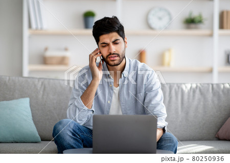 Remote business. Concerned arab freelancer talking on cellphone and using laptop computer, sitting on sofa at home 80250936
