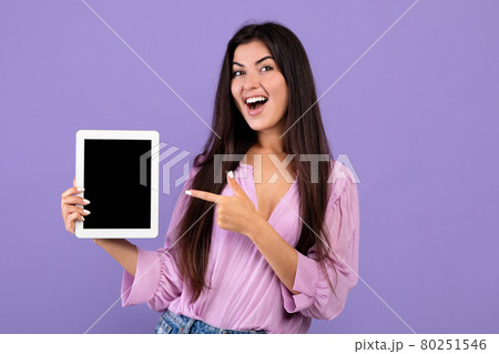 Check this.Cheerful armenian woman holding and pointing at digital tablet with black blank screen, purple background 80251546