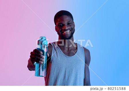 Smiling young black sportsman showing bottle with fresh water in neon light. Fitness, active lifestyle, wellness concept Smiling young black sportsman showing bottle with fresh water in neon light. Fitness, active lifestyle, wellness concept 80251588
