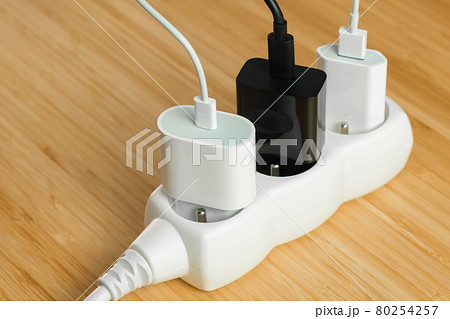 The electrical extension strip with connected white and black power plugs on the wooden table. Smart phones chargers in power strip for electrical sockets The electrical extension strip with connected white and black power plugs on the wooden table. Smart phones chargers in power strip for electrical sockets 80254257