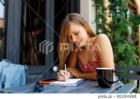 Young woman on cafe terrace. Hipster stylish concentrated girl Writing In Notebook. Digital technologies For Remote Working or studying at comfortable place, coffee or tea drinking. Young woman on cafe terrace. Hipster stylish concentrated girl Writing In Notebook. Digital technologies For Remote Working or studying at comfortable place, coffee or tea drinking. 80264988