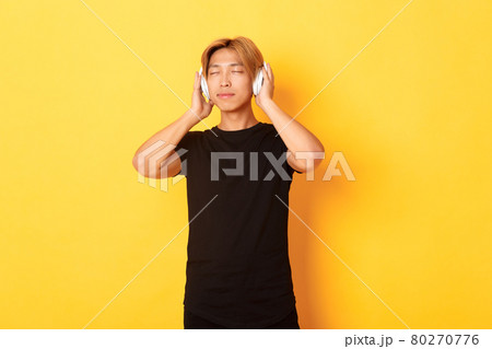 Relaxed and pleased handsome asian guy close eyes from satisfaction while listening music in wireless headphones, standing yellow background Relaxed and pleased handsome asian guy close eyes from satisfaction while listening music in wireless headphones, standing yellow background 80270776