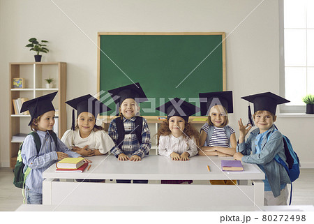 Group of happy school kids in academic caps standing by table with books in classroom Group of happy school kids in academic caps standing by table with books in classroom 80272498