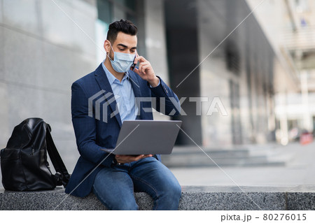 Arab CEO in face mask with laptop having phone conversation Arab CEO in face mask with laptop having phone conversation 80276015